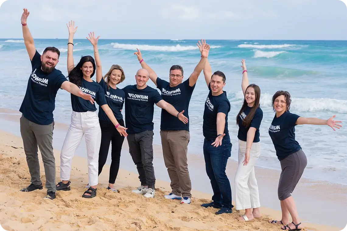 Flowium team group photo on a sandy beach with ocean in the background, eight members wearing matching black t-shirts with white logo, posing with raised arms in a cheerful moment.
