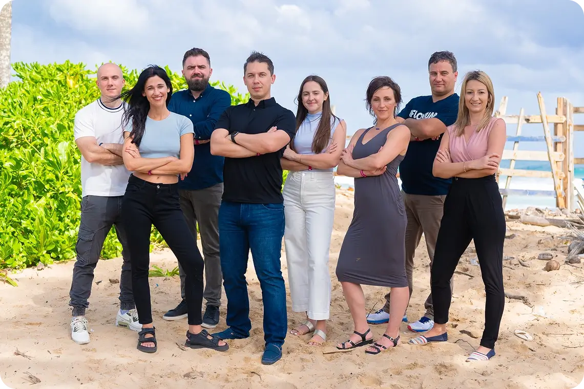 Flowium team group photo on a sandy beach with ocean in the distance, eight members standing side by side with arms crossed, posing together in front of greenery and a wooden fence.