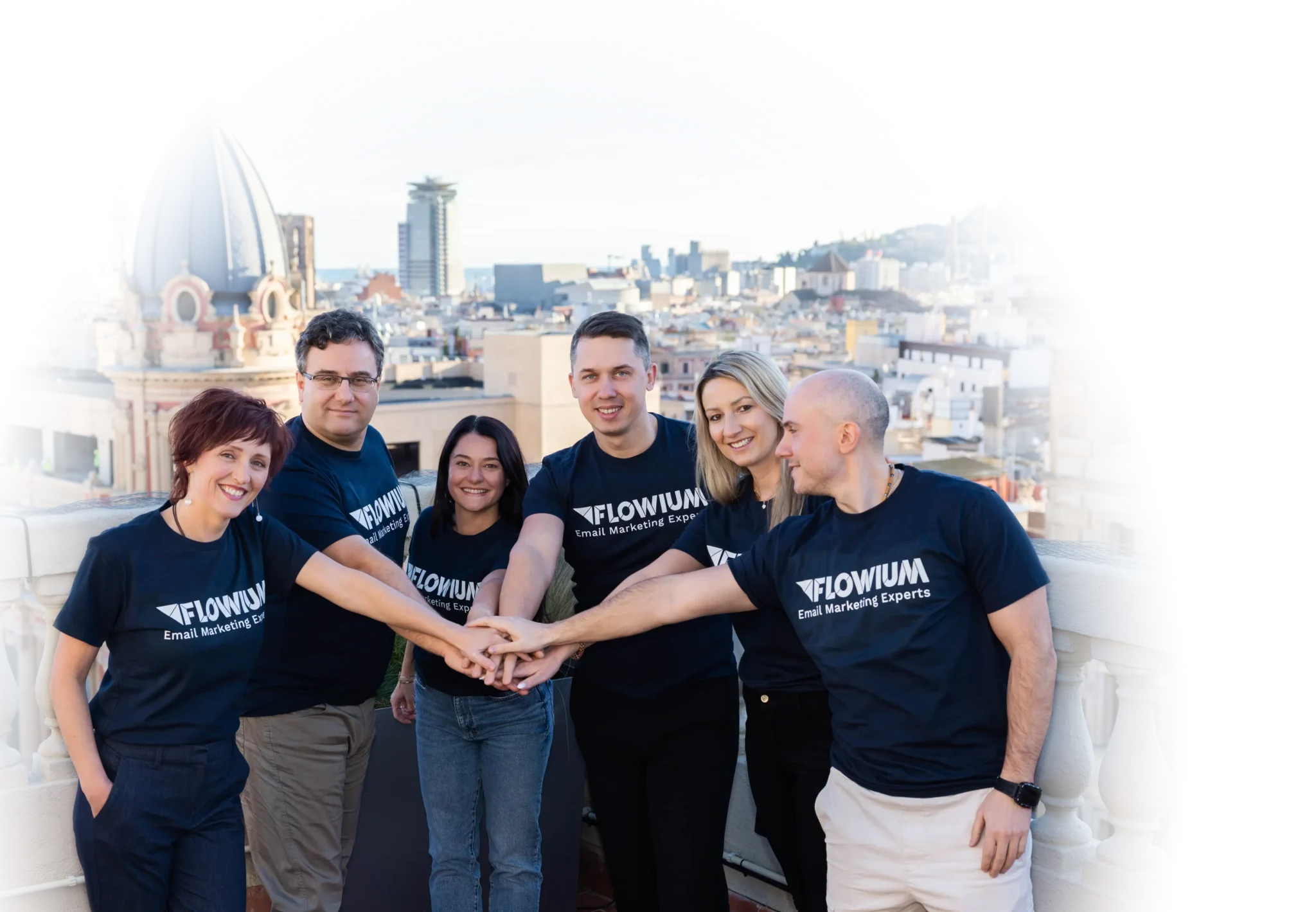 A group of six Flowium team members stand on a rooftop in front of a cityscape, wearing matching navy t-shirts with the logo “FLOWIUM Email Marketing Experts.” They form a semi-circle with their hands stacked together in the center, symbolizing teamwork and unity